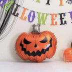 An orange jack-o'-lantern foil balloon with a black face and stem, displayed on a white surface against a white wall with a colorful Halloween banner in the background.